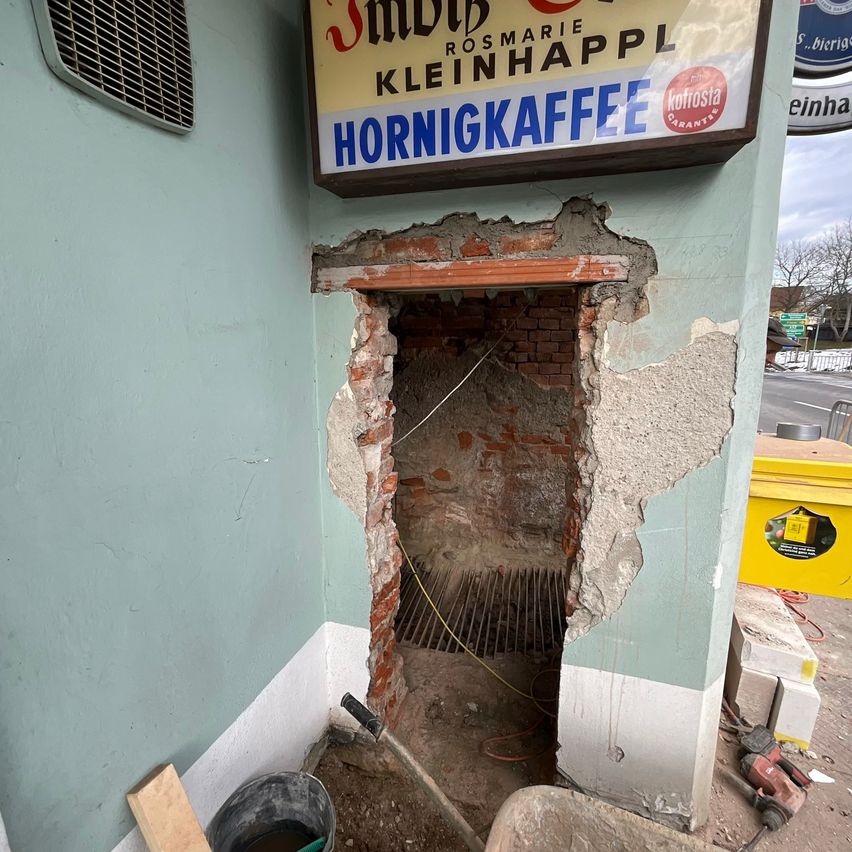 A damaged brick fireplace with a sign for Hornigkaffee on a green wall. Nearby, a bucket and tools are present, indicating ongoing construction.