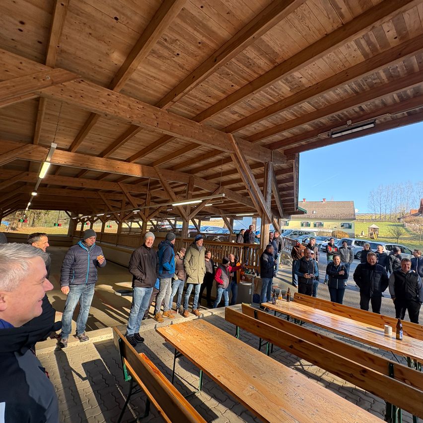 Eine Gruppe von Menschen versammelt sich in einer hölzernen Außenstruktur mit Blick auf einen Parkplatz. Die Struktur hat Holzbalken und einen Holzboden. Einige Leute stehen, während andere auf Bänken sitzen. Es gibt aufgestellte Tische und Stühle.