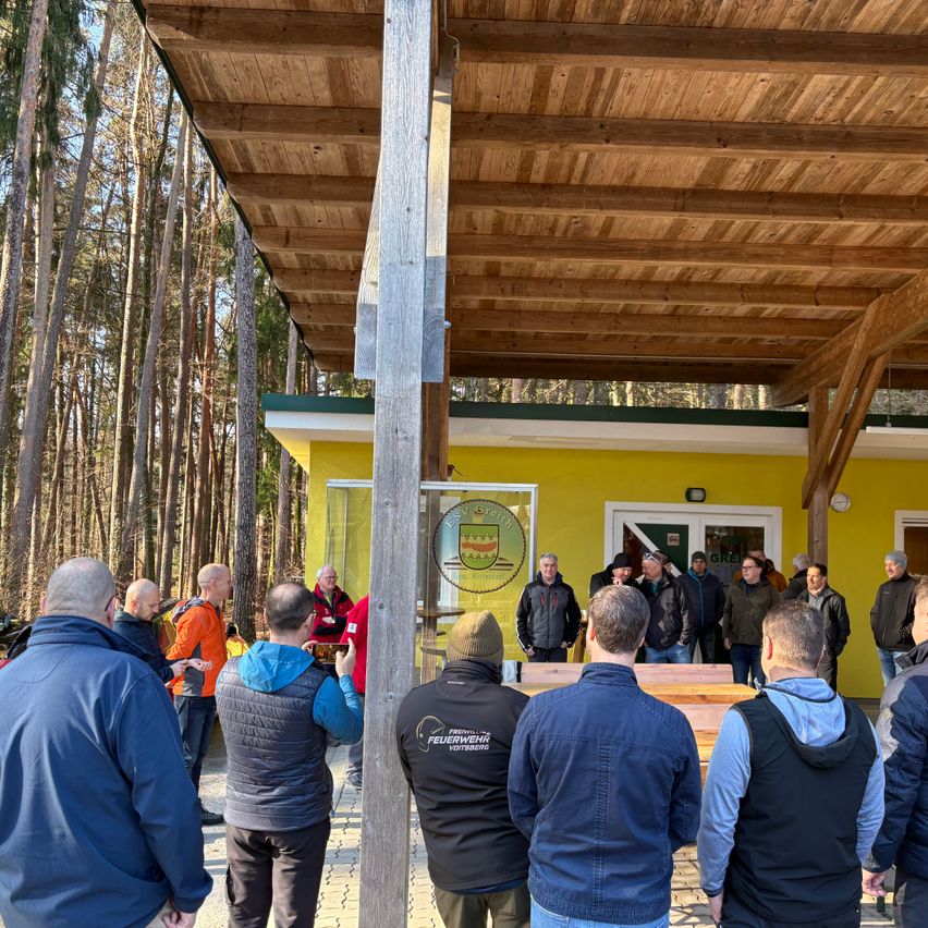 Eine Gruppe von Menschen, in Winterkleidung gekleidet, versammelt sich unter einem hölzernen Schutzdach in einem Wald. Ein gelbes Gebäude ist im Hintergrund zu sehen.