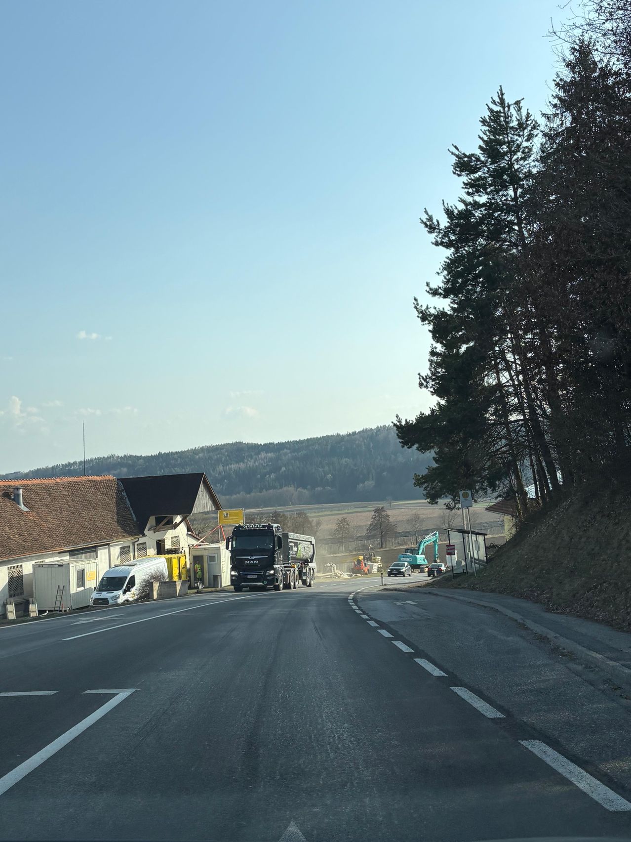 Eine Straße mit einem geparkten Lkw und einem Van, Bäumen und Gebäuden auf der linken Seite und Maschinen auf der rechten Seite. Ein Gebirgszug in der Ferne.
