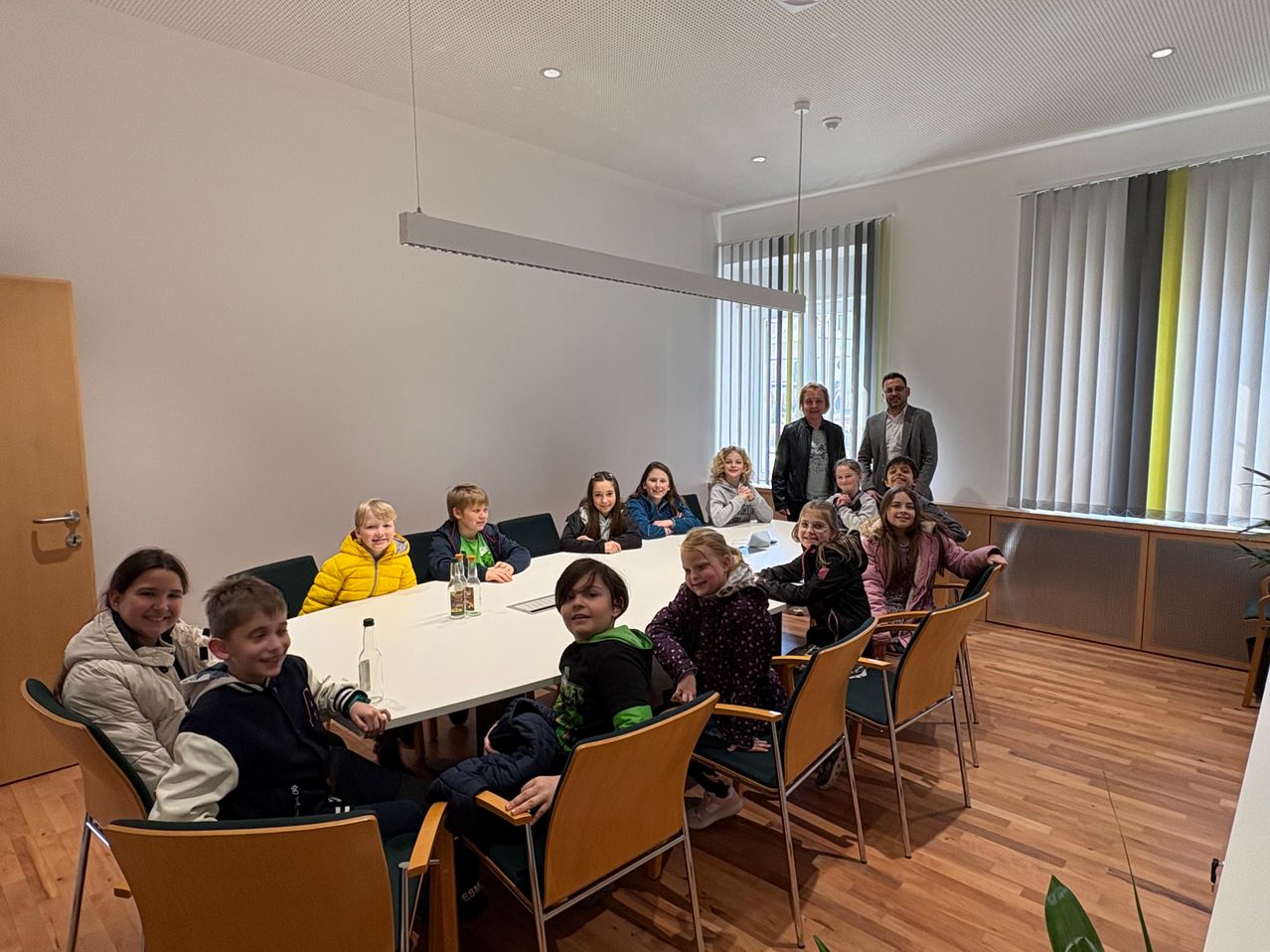 Eine Gruppe von Kindern und einige Erwachsene sitzen um einen Tisch in einem Konferenzraum. Flaschen stehen auf dem Tisch, und zwei Erwachsene stehen hinter den sitzenden Personen.