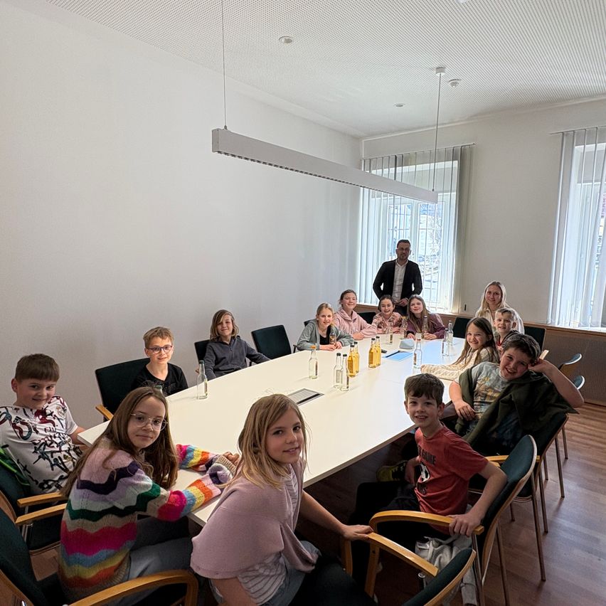 Eine Gruppe von Kindern und einem Mann sitzen um einen langen Tisch in einem Raum. Sie lächeln, mit Flaschen und einem Laptop auf dem Tisch. Eine Frau steht hinter dem Tisch.