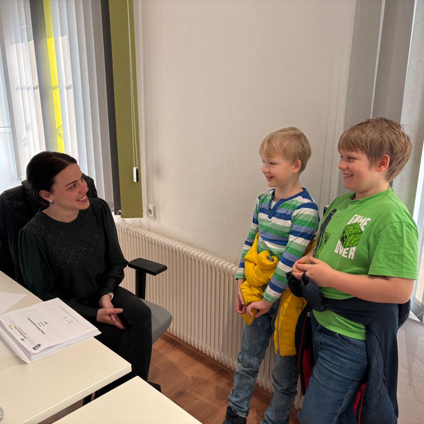 Eine Frau lächelt, während sie in einem Stuhl sitzt, mit zwei Jungen neben ihr, einer in einer gelben Jacke und der andere in einem grünen Minecraft-T-Shirt. Sie befinden sich in einem Raum mit einer weißen Wand, einem Heizkörper und einem Tisch mit Papieren.