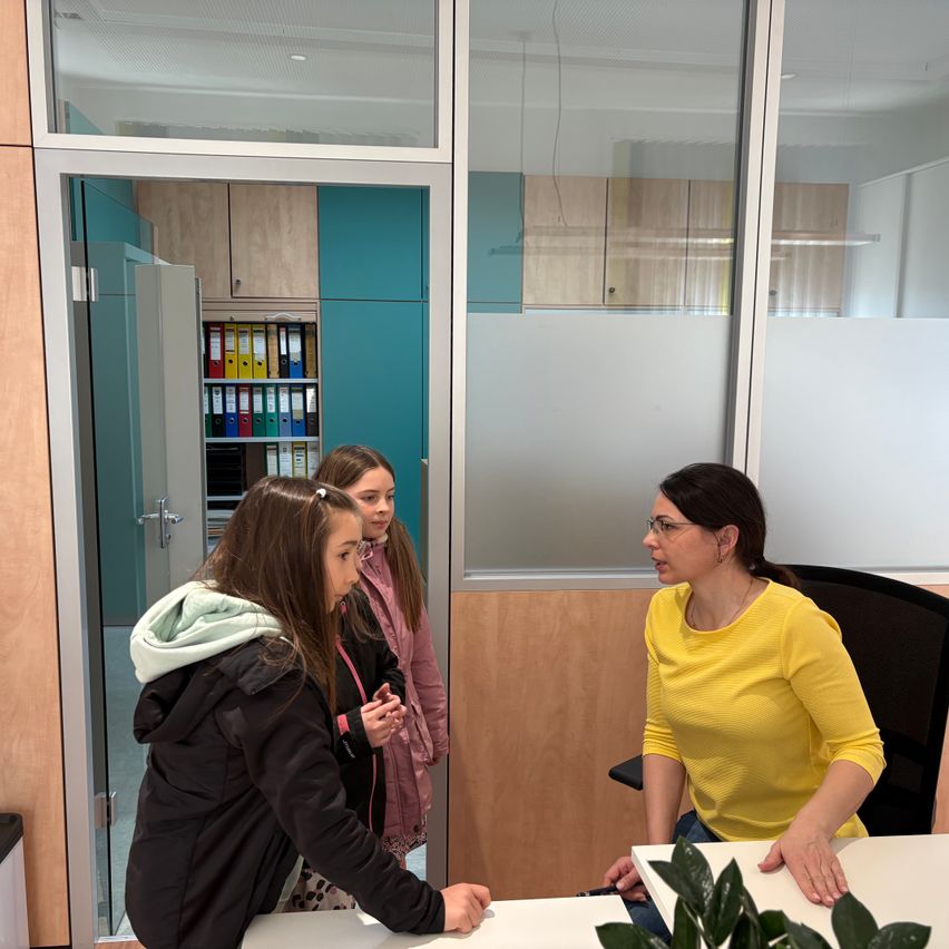 Drei junge Mädchen sind in einem Büroraum. Zwei Mädchen stehen an einer Glastür, während das dritte Mädchen an einem Schreibtisch sitzt und mit ihnen spricht.