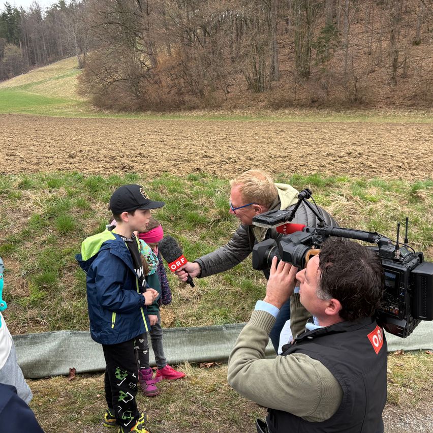 Ein Mann mit einer Kamera interviewt einen Jungen in einer blauen Jacke und ein Mädchen in einer rosa Mütze auf einem Feld.