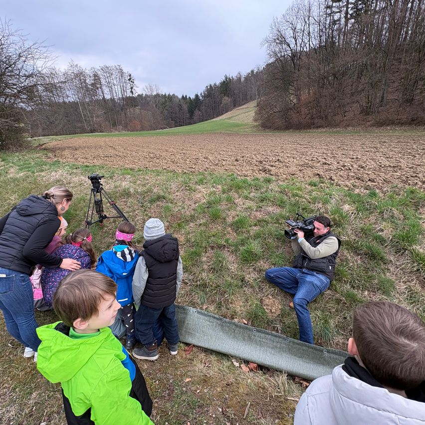 Eine Gruppe von Kindern und Erwachsenen steht auf einem Grasfeld, einige beobachten einen Mann, der mit einer Kamera auf einem Stativ filmt.