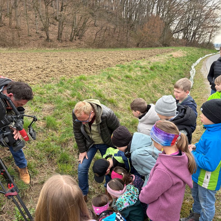 Eine Gruppe von Kindern und Erwachsenen versammelt sich auf einem Grasfeld mit Bäumen im Hintergrund. Ein Mann hält ein Mikrofon und eine Kamera.