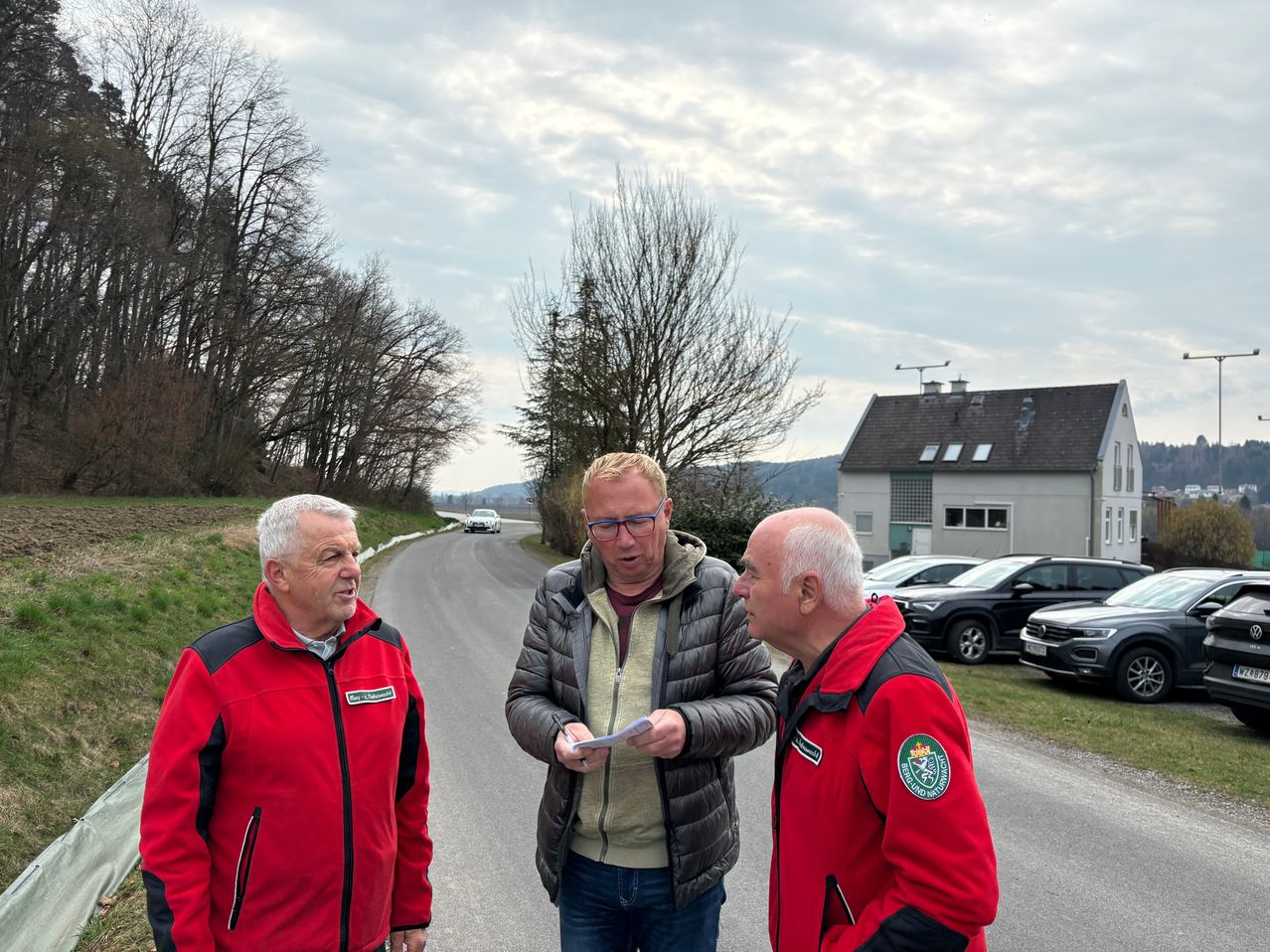 Drei Männer in Jacken stehen auf einer Straße, einer hält ein Papier. Hinter ihnen fährt ein Auto die Straße hinunter, und ein Haus mit geparkten Autos ist sichtbar.
