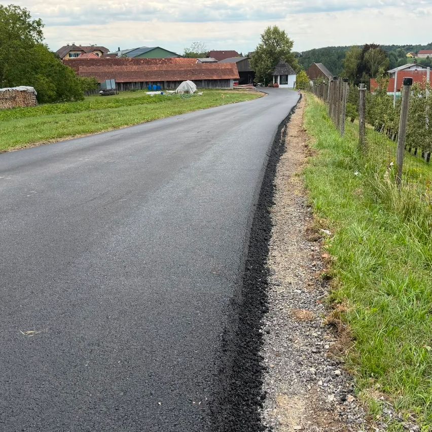 Eine neu asphaltierte Straße mit einem Schotterrand und einem Grasrand, mit Häusern und Bäumen im Hintergrund.