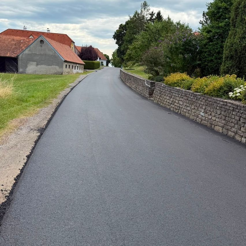 Eine neu asphaltierte Straße mit einer Steinmauer auf einer Seite und einem Grasbereich auf der anderen, die zu einem Haus mit rotem Dach führt.