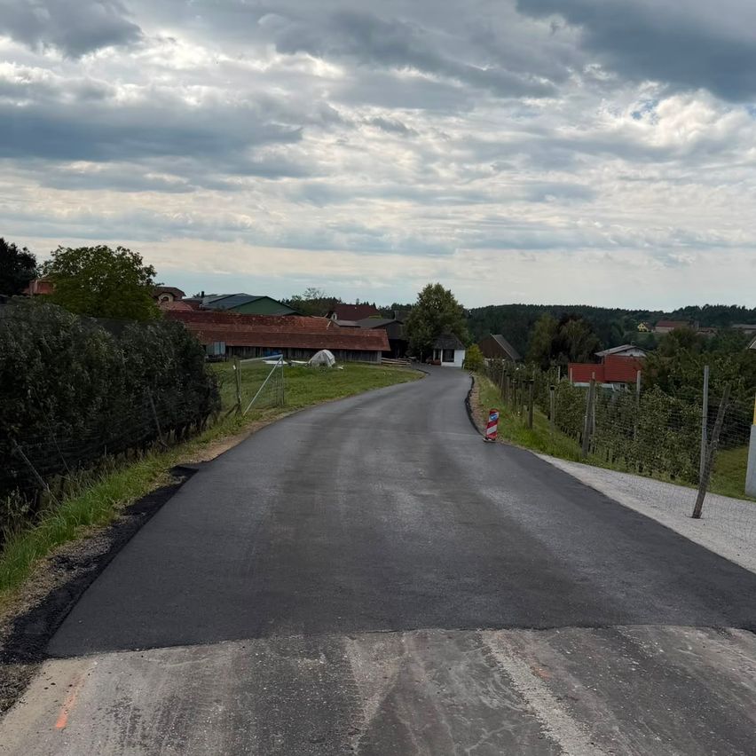 Eine frisch asphaltierte Straße in einem ländlichen Gebiet mit Häusern und Grünflächen auf beiden Seiten unter einem bewölkten Himmel.