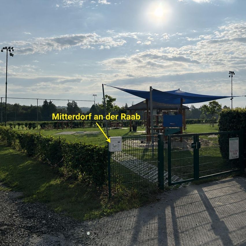 Ein sonniger Tag in einem Park in Mitterdof an der Raab, mit einem blauen Vordach, einem grünen Zaun und einem gelben Schild, das auf den Eingang weist.