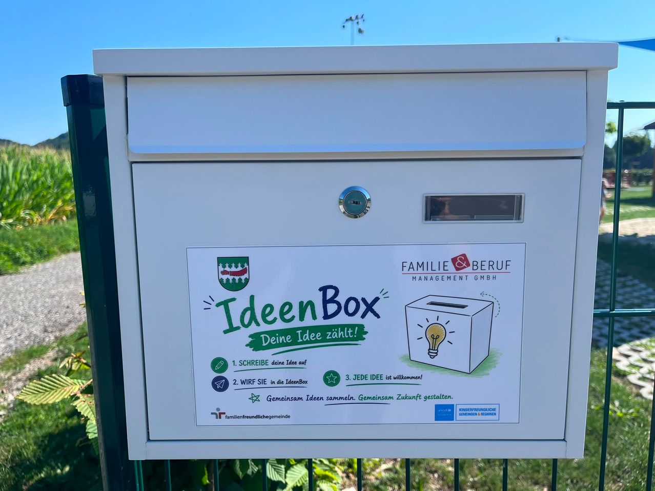 Eine weiße IdeenBox mit einer Lichtbirne-Zeichnung und Anweisungen zum Einreichen von Ideen. Die Box ist an einem grünen Zaun montiert.