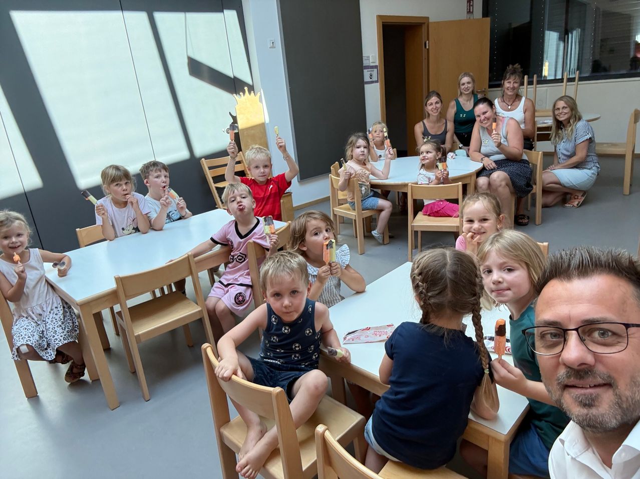 A group of children and adults are sitting around tables, eating ice cream in a room with large windows.