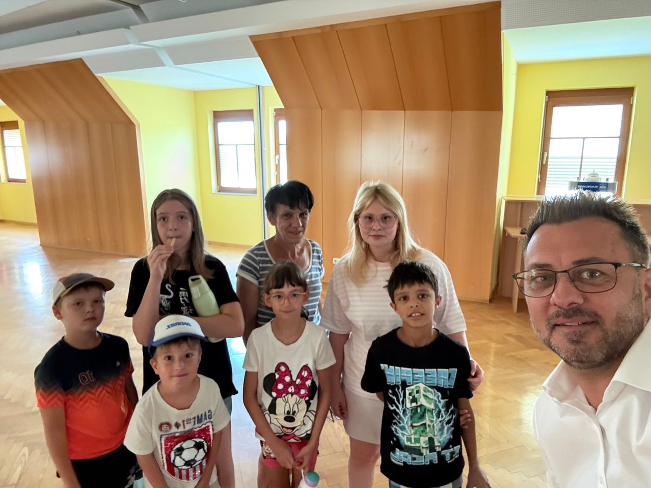 A group of children and adults stand in a room with yellow walls, smiling for a photo. The adults include a woman in glasses and a man with a beard. The children wear various t-shirts, one with a soccer ball design.
