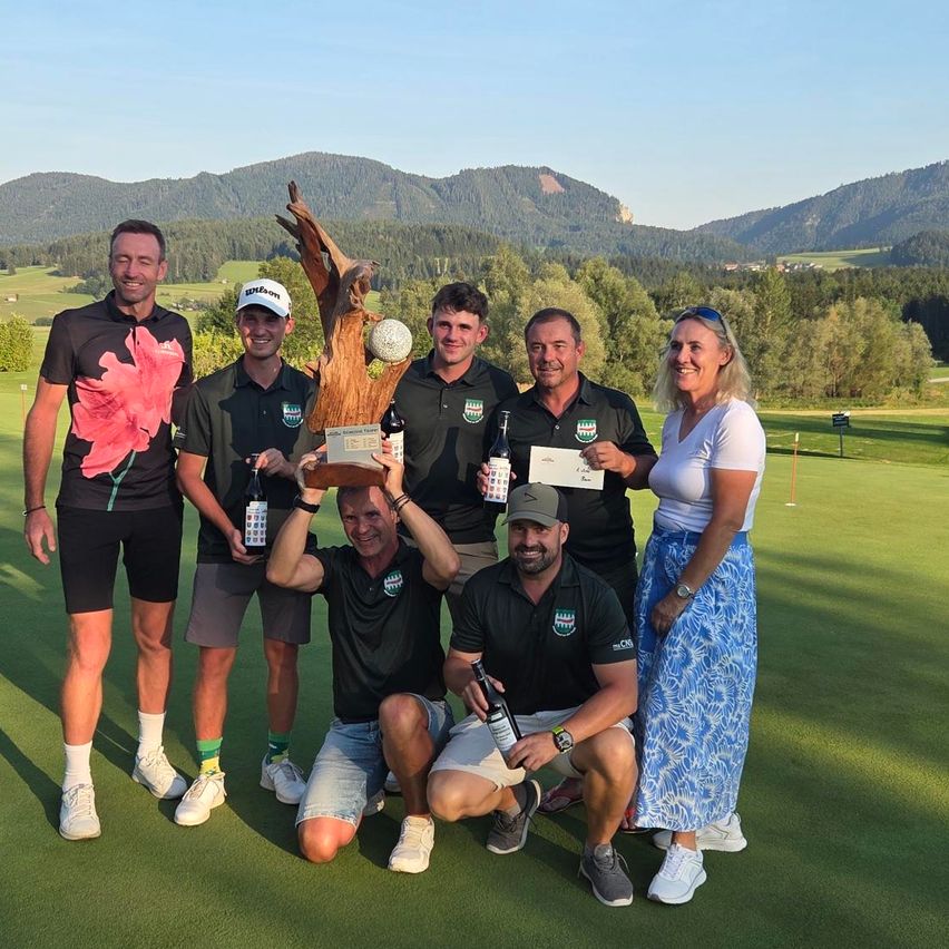 A group of golfers, both men and women, are posing for a photo on a golf course with mountains in the background. They are holding trophies, bottles, and certificates.