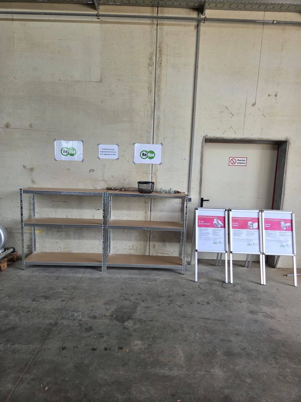 An empty storage room with metal shelves and a basket on top. Three posters are on the wall, and a door is on the right.
