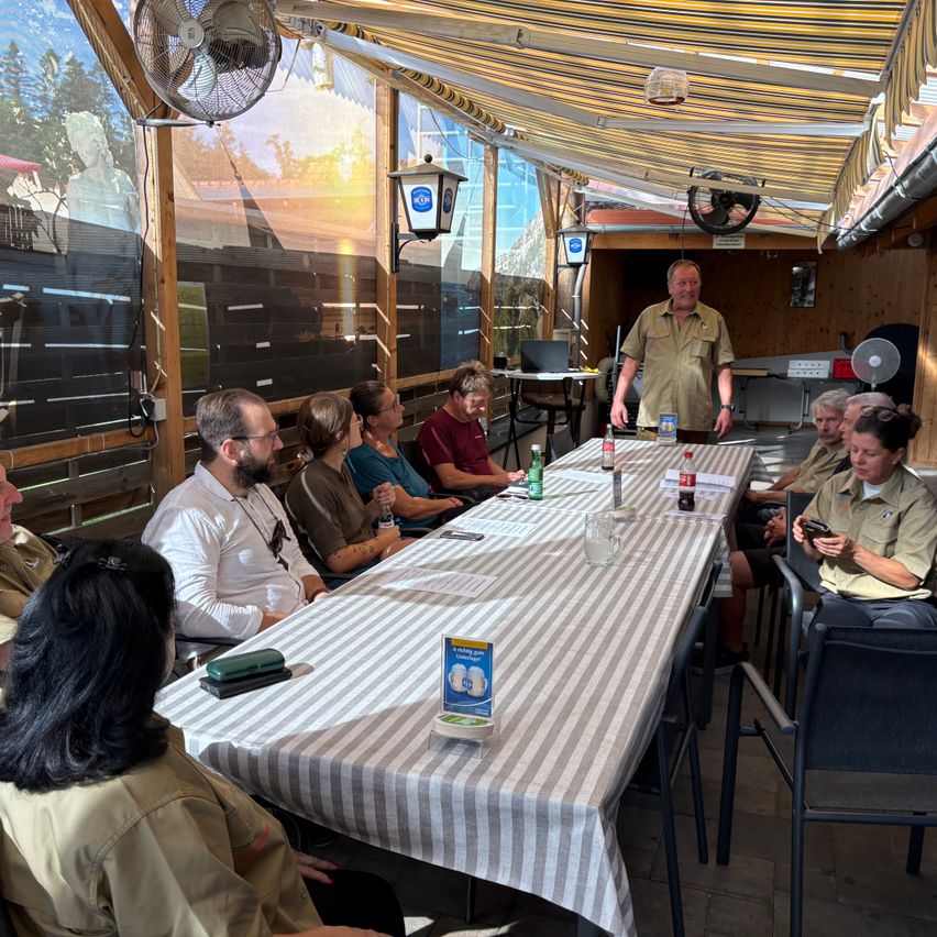 A group of people sit around a long table in an outdoor restaurant. A man stands at the head of the table, and a box is on the table.