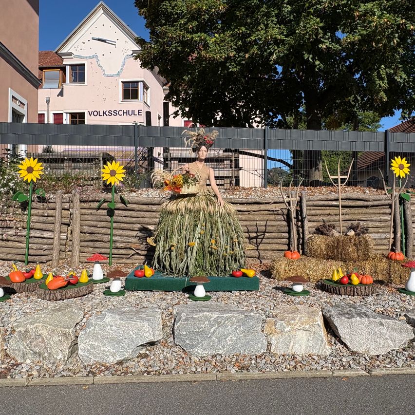 A garden display featuring a woman statue dressed in a grass skirt, holding a basket of flowers, surrounded by pumpkins, pears, and mushrooms on a gravel path. Behind her is a wooden fence with sunflowers and a building with the sign 'Volksschule'.