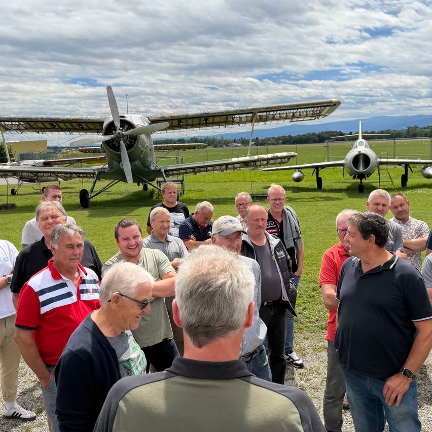 Eine Gruppe von Menschen steht auf einem Rasenfeld neben zwei Flugzeugen, eines davon ein altes Doppeldecker, unter einem teilweise bewölkten Himmel.