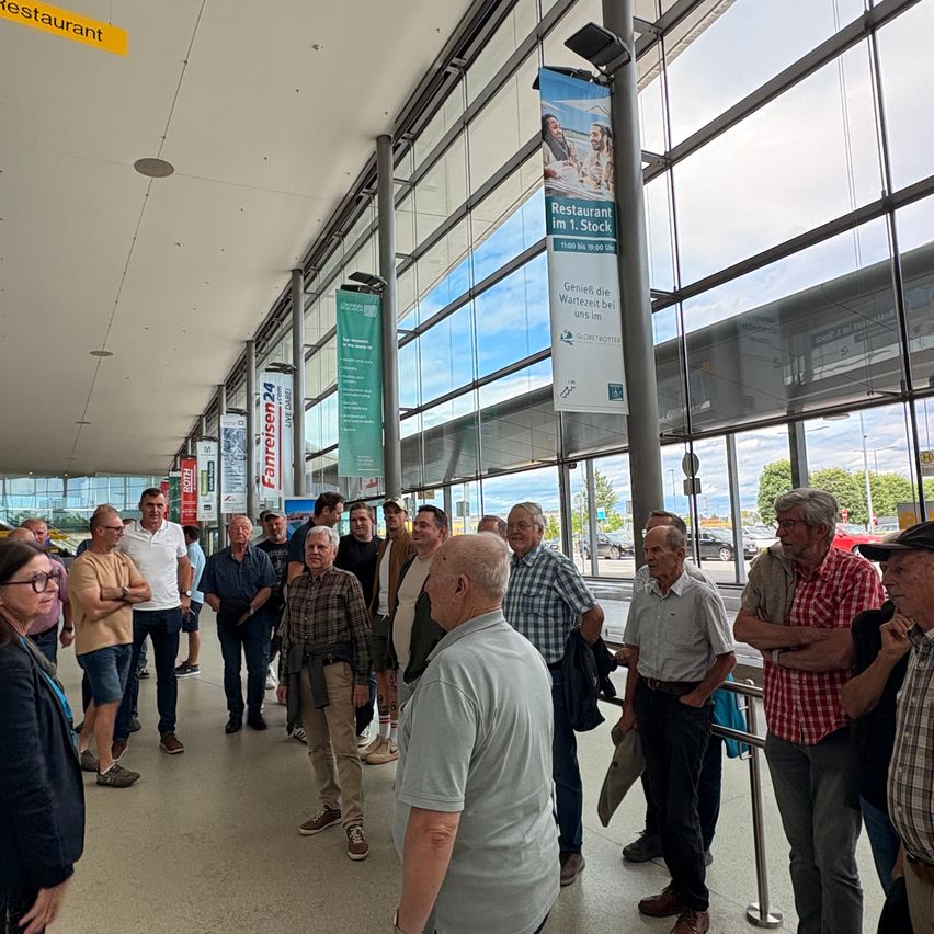 Eine Gruppe von Menschen steht in einer Schlange an einem Flughafen, wahrscheinlich im Wartemodus. Sie schauen sich etwas an, wahrscheinlich ein Ankündigungsboard. Der Ort hat Glaswände und eine Decke. Es hängen auch Banner an den Glaswänden. Ein Restaurant-Schild ist an der Decke. Draußen sind Autos geparkt.