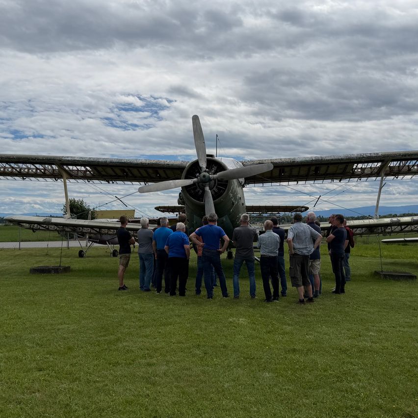 Eine Gruppe von Menschen steht vor einem Oldtimer-Flugzeug mit Propeller auf Gras unter einem bewölkten Himmel.