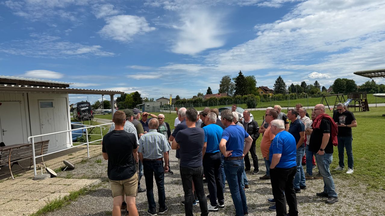 Eine Gruppe von Männern steht in einem Kreis auf einem Kiesfeld. Sie tragen Freizeitkleidung, und einige von ihnen tragen Sonnenbrillen. Hinter ihnen befindet sich ein Rasenbereich mit einem weißen Zaun. Bäume und Gebäude sind in der Ferne zu sehen, und der Himmel ist blau mit Wolken.