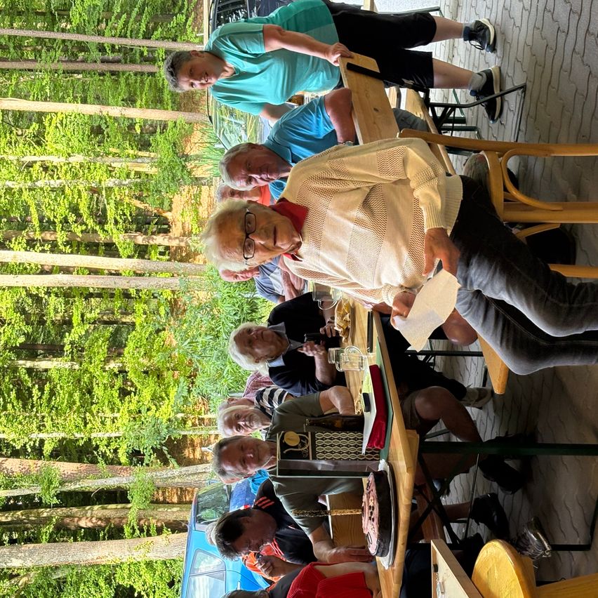 Eine Gruppe von Menschen sitzt auf Stühlen um einen Tisch in einem Park. Sie scheinen ein Picknick zu machen. Einige von ihnen halten Tassen. Der Tisch ist mit einem roten Tuch bedeckt.