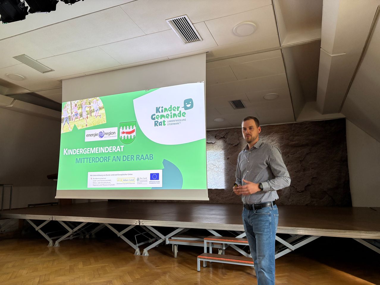 Ein Mann steht vor einer Projektionsleinwand mit einer Präsentation mit dem Titel 'Kindergemeinderat Mitterdorf an der Raab'. Er trägt ein Hemd und Jeans. Hinter ihm zeigt eine große Projektionsleinwand eine grüne und weiße Präsentation.