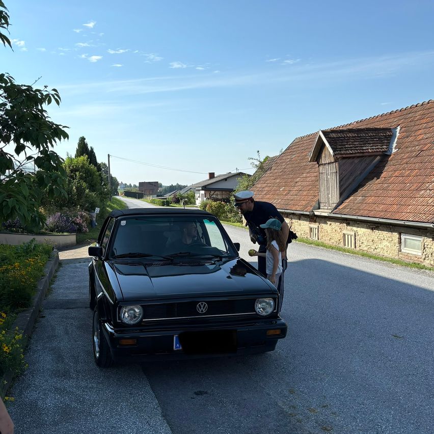 A black Volkswagen Golf with a license plate is parked on the side of the road with a man and a child inspecting it.