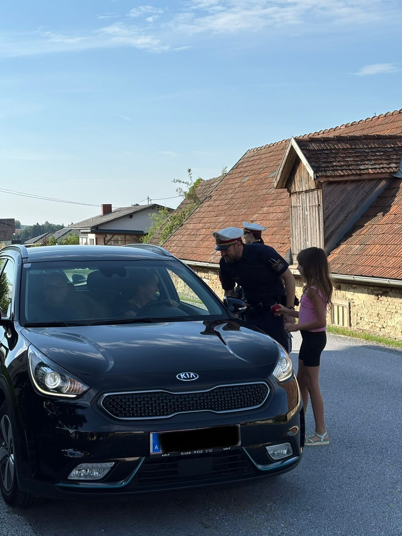 A police officer checks a car, with a young girl handing him an apple. The car has a Kia logo on the front. Behind it are two houses with brown roofs.