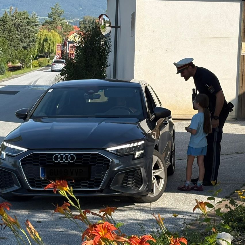 A policeman speaks with a girl next to a black Audi car in front of a building. Trees and a car on the road are in the background.