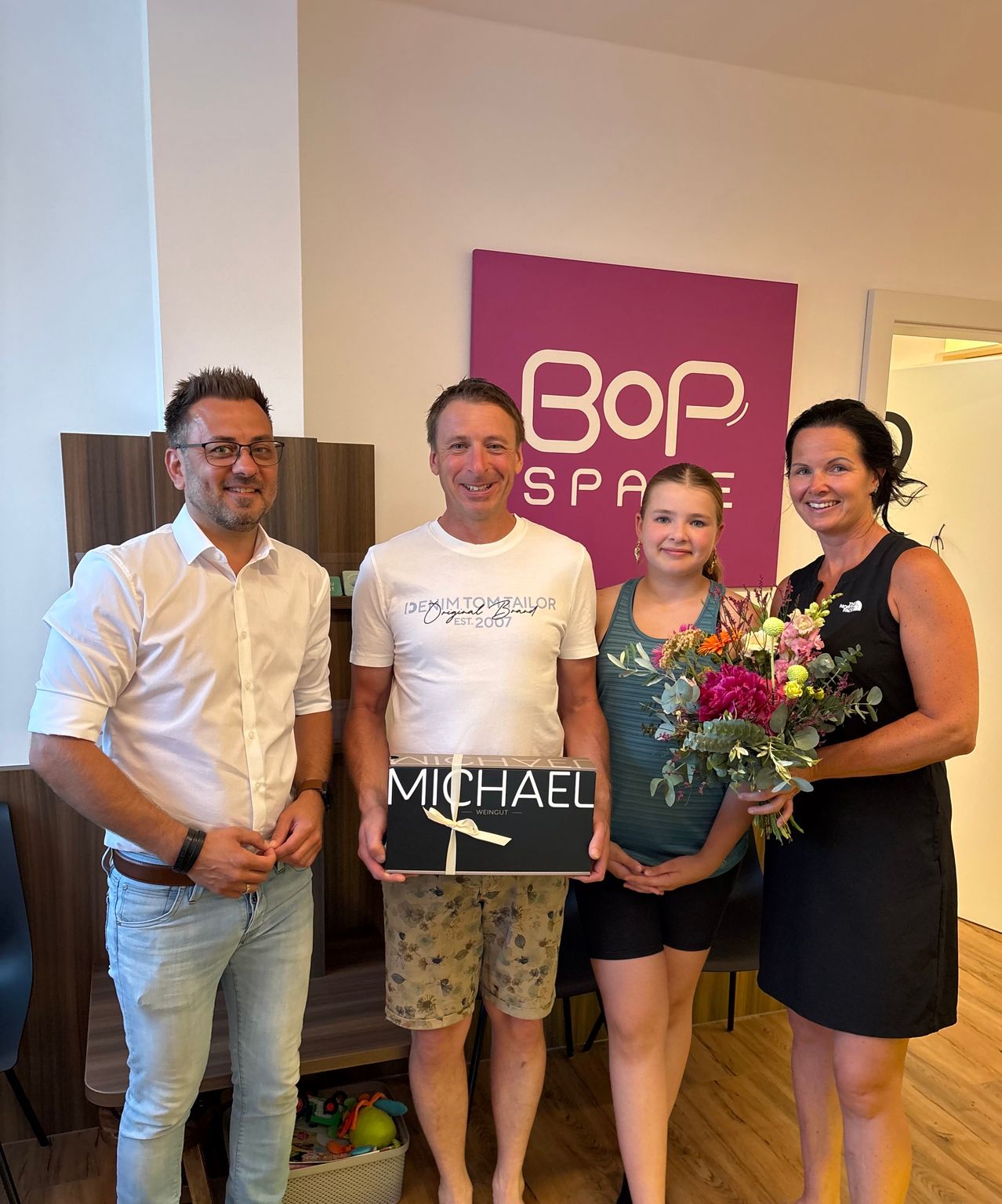 Three people are posing for a photo with a sign in the background. One person is holding a flower bouquet, another a gift box with the name Michael.