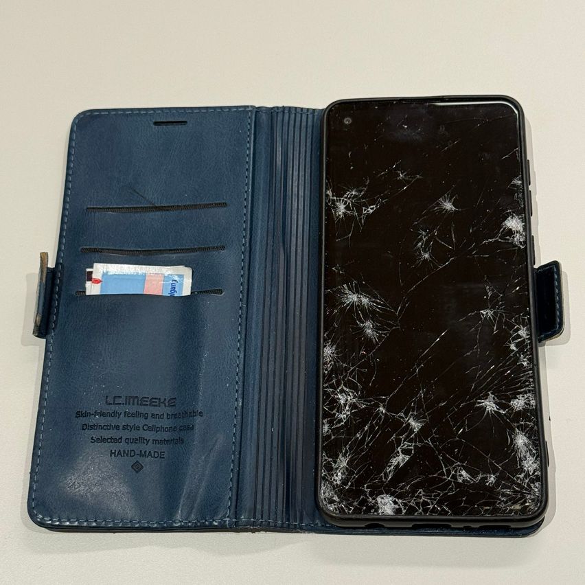 An open blue smartphone case with a cracked screen, placed on a white surface. The case has a wallet design and includes a few cards.