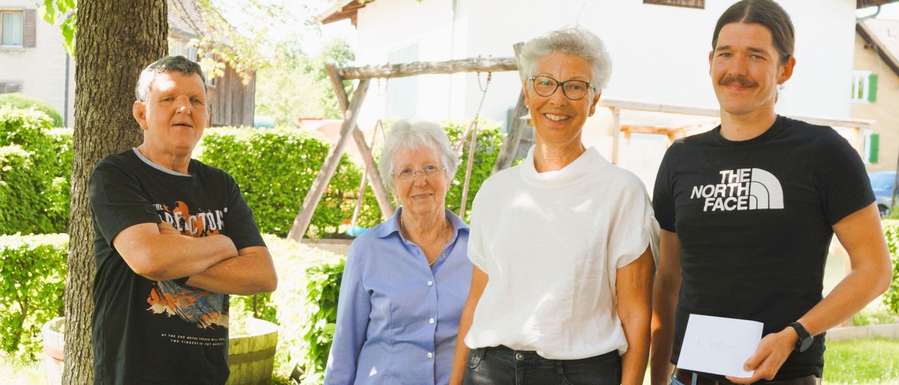 Vier Personen stehen lächelnd in einem Garten unter einem Baum für die Kamera. Ein Mann links hat die Arme verschränkt, während ein Mann rechts ein Stück Papier hält. Dahinter trägt eine Frau eine Brille und eine Halskette.
