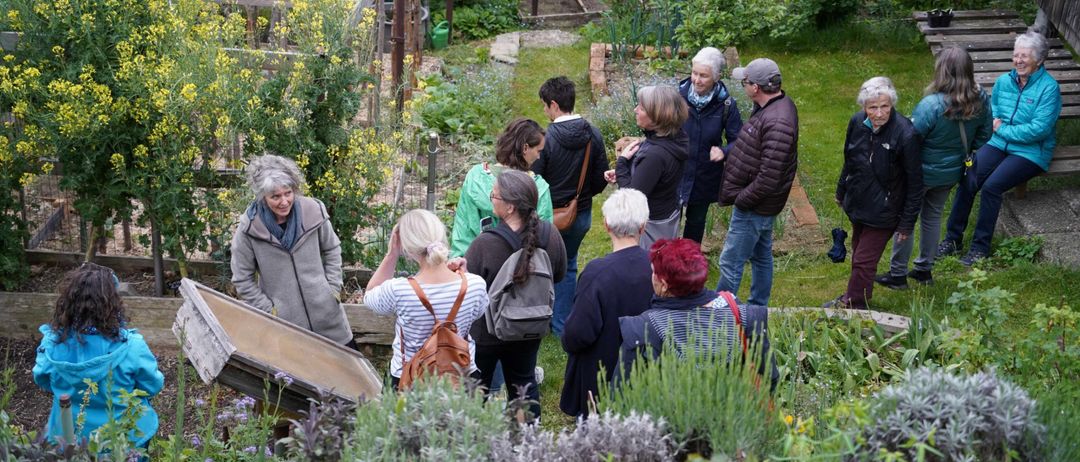 Eine Gruppe von Menschen versammelt sich in einem Garten, diskutiert und betrachtet Pflanzen. Einige tragen Jacken und Mützen, und im Hintergrund befindet sich eine Holzstruktur.