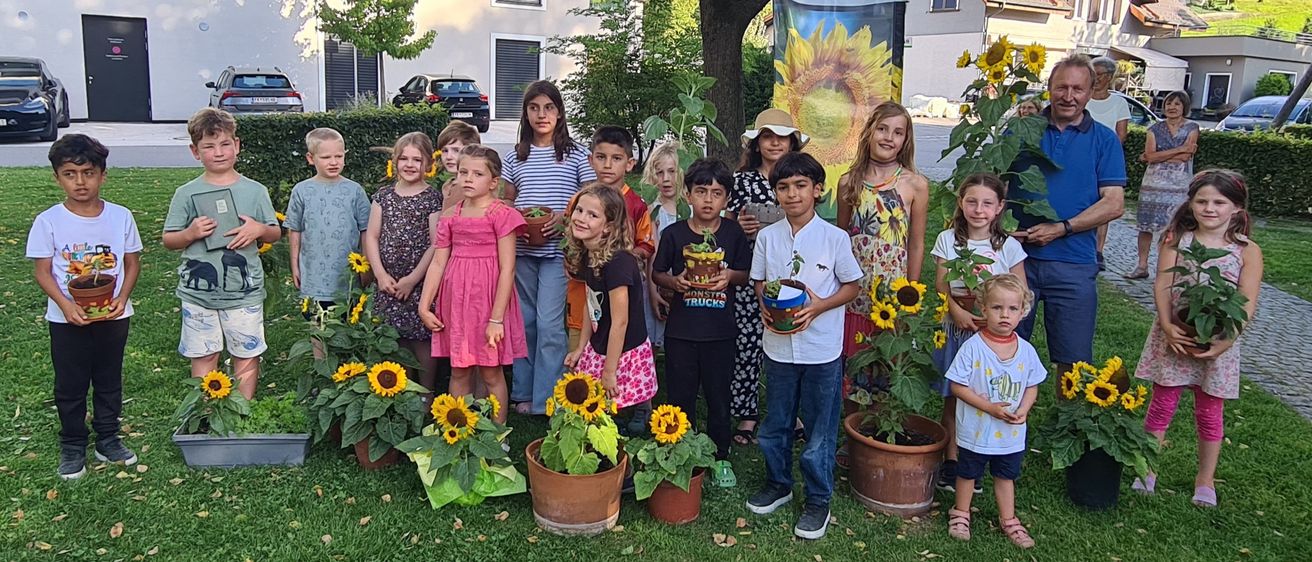 Eine Gruppe von Kindern und Erwachsenen posiert für ein Foto in einem Garten mit Sonnenblumen und einem Banner im Hintergrund.