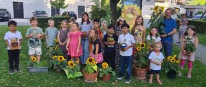 Eine Gruppe von Kindern und Erwachsenen posiert für ein Foto in einem Garten mit Sonnenblumen und einem Banner im Hintergrund.