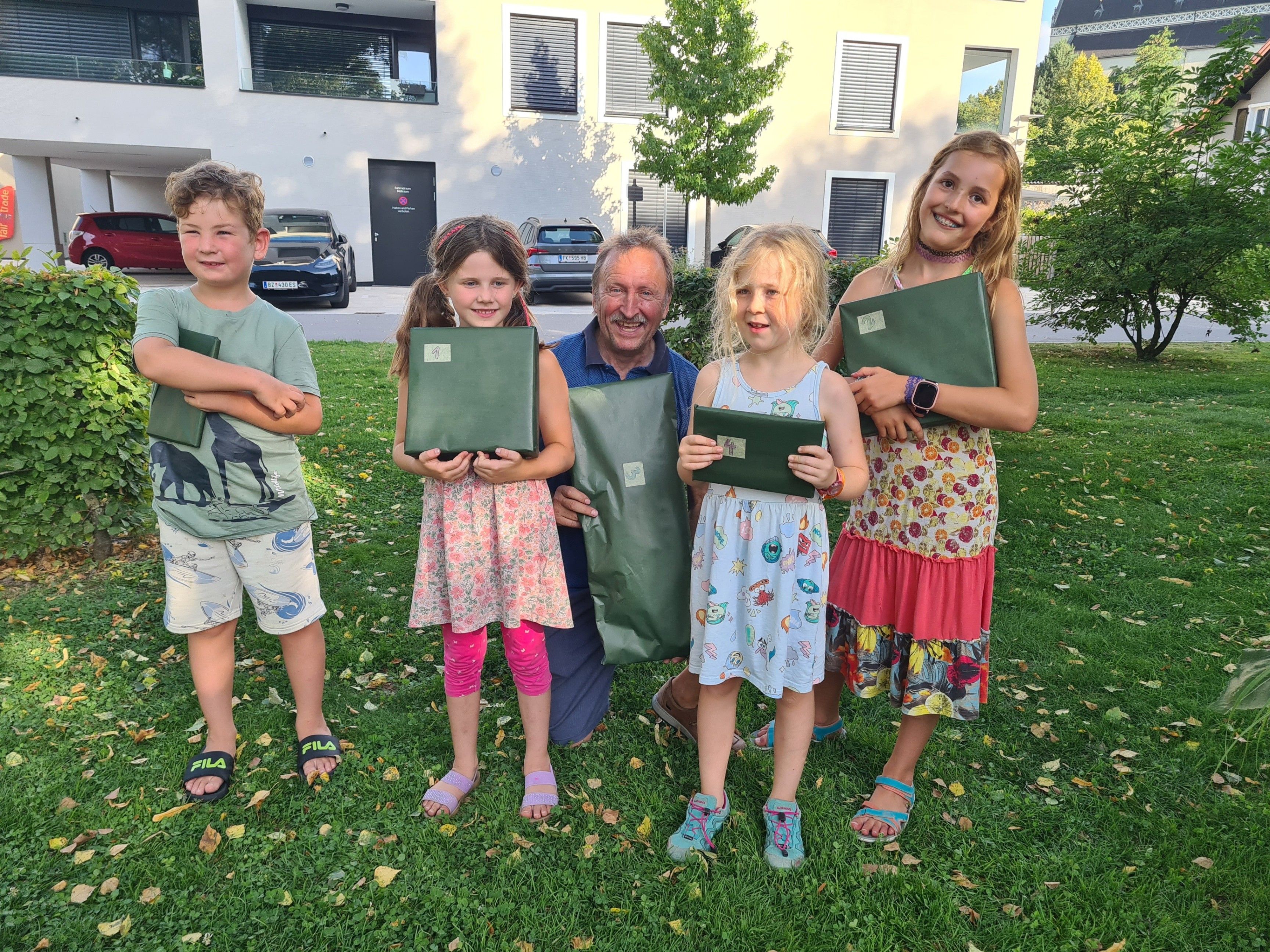 Eine Familie steht auf dem Rasen, ein Mann kniet, alle halten grüne Ordner. Hinter ihnen steht ein Gebäude mit geparkten Autos und einem Baum.