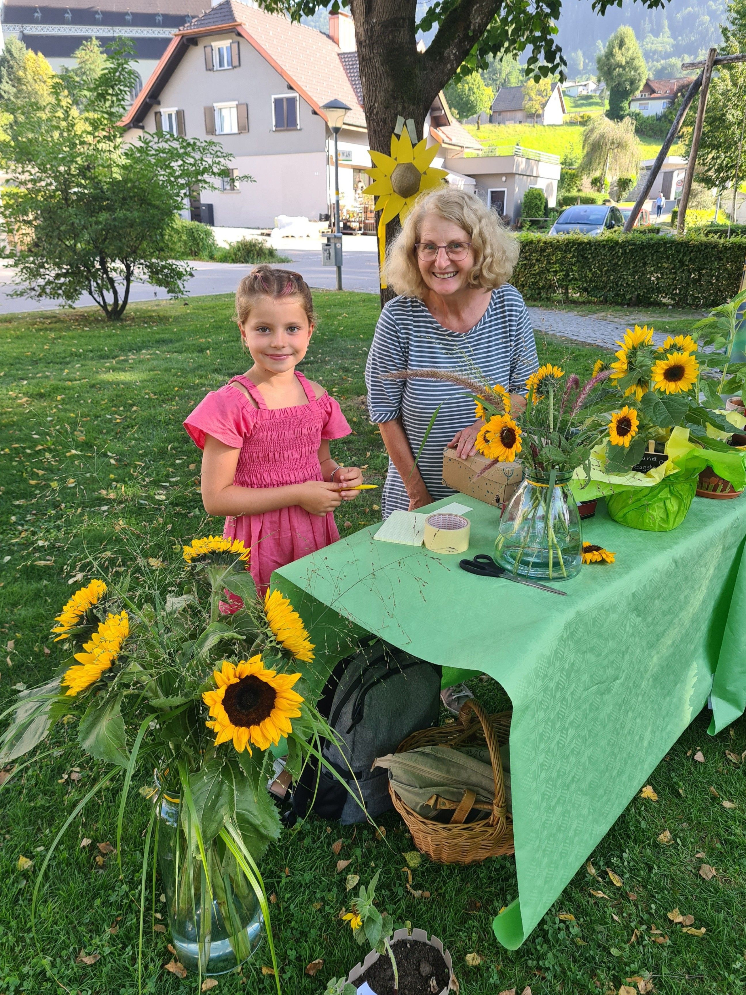 Eine Frau und ein junges Mädchen stehen hinter einem Tisch mit Sonnenblumen in einer Vase. Sie lächeln und scheinen sich auf einer Veranstaltung zu befinden.
