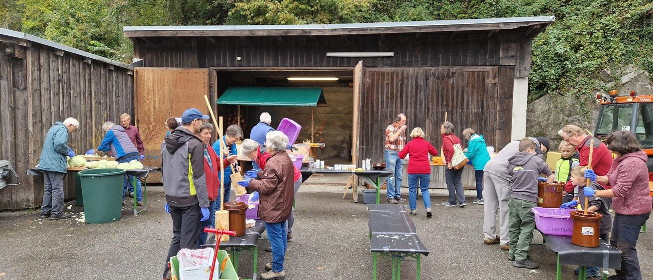 Mehrere Personen stehen vor einem Gebäude, wahrscheinlich bei einer Outdoor-Aktivität. Einige von ihnen tragen Handschuhe und scheinen an etwas zu arbeiten. Draußen stehen Tische und Bänke. Ein Trolley befindet sich auf der linken Seite, und einige Eimer sind auf dem Tisch platziert.