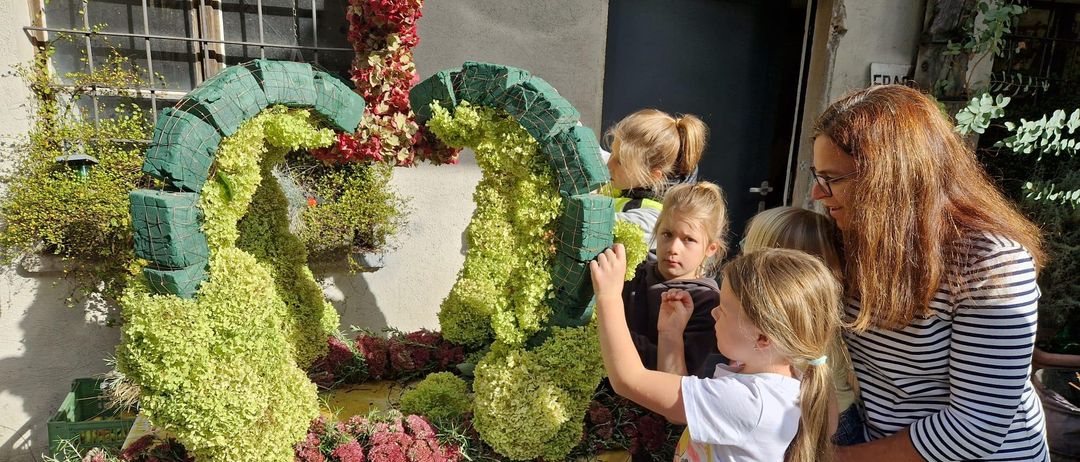 Eine Gruppe von Kindern berührt eine aufwendige Blumenarrangement auf einem Tisch draußen. Es besteht aus grünen und roten Pflanzen, mit einer Frau in der Nähe.