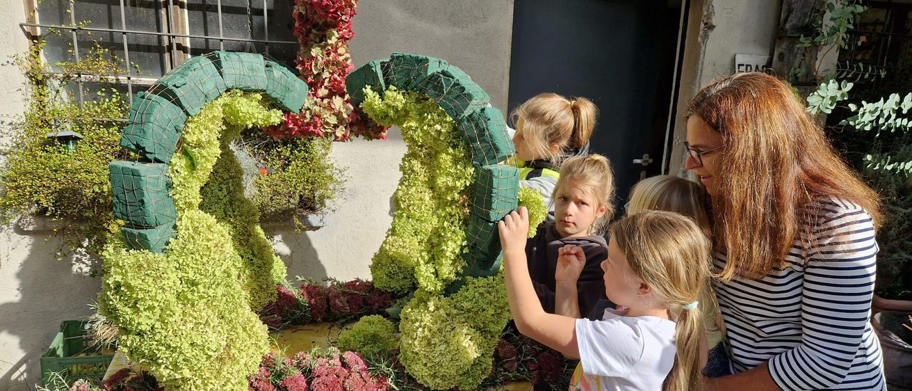 Eine Gruppe von Kindern berührt eine aufwendige Blumenarrangement auf einem Tisch draußen. Es besteht aus grünen und roten Pflanzen, mit einer Frau in der Nähe.