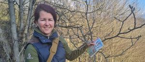 Eine lächelnde Frau im Freien hält einen Ast und ein Handbuch und steht unter einem kahlen Baum.