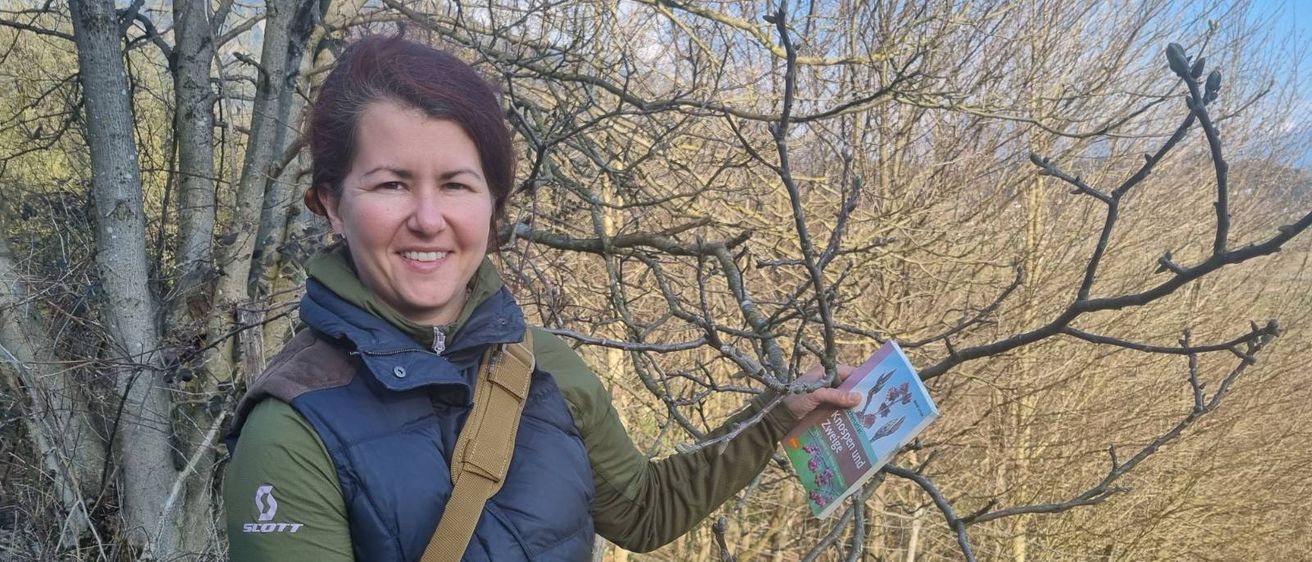 Eine lächelnde Frau im Freien hält einen Ast und ein Handbuch und steht unter einem kahlen Baum.