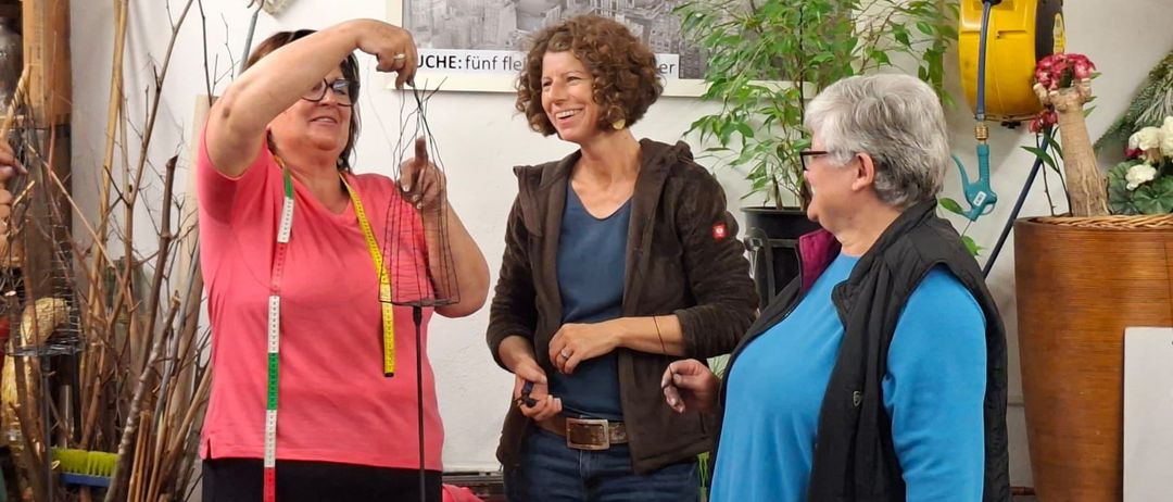 Drei Frauen stehen in einer Werkstatt. Eine hält ein Maßband, eine andere lächelt. An der Wand hinter ihnen ist ein Foto von Arbeitern zu sehen. Werkzeuge und eine Topfpflanze sind in der Nähe.