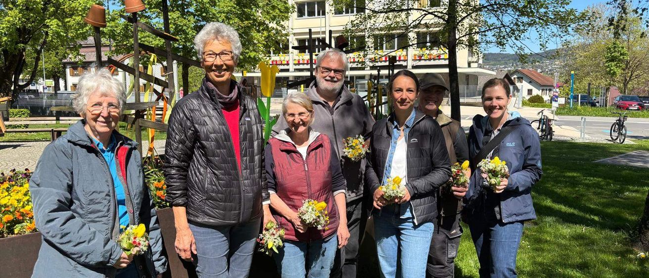 Eine Gruppe von Menschen steht in einem Park und hält Blumen in der Hand. Hinter ihnen befindet sich ein Gebäude und ein Baum. Eine Frau trägt eine Brille und ein Halstuch. Sie scheinen für ein Foto zu posieren.