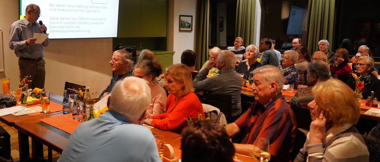 Eine Gruppe älterer Erwachsener sitzt in einem Konferenzraum um einen Tisch herum und hört einer Präsentation auf einer Projektionsleinwand zu. Der Raum hat grüne Vorhänge und mehrere Fenster.