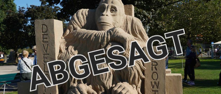 Eine Sandplastik eines Gorillas sitzt auf drei Steinblöcken, mit den Wörtern 'ABGESAGT' und 'WACHSEN' eingraviert. Die Skulptur befindet sich im Freien mit Bäumen im Hintergrund.