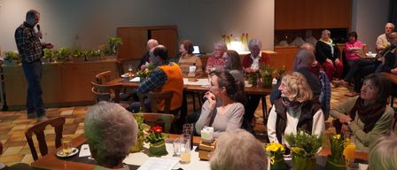 Eine Gruppe von Menschen, überwiegend ältere Erwachsene, sitzt in einem Esszimmer um Tische herum, möglicherweise bei einem Treffen. Jeder Tisch hat Blumen, Gläser und Papiere. Der Raum hat weiße Wände und Holzschränke.